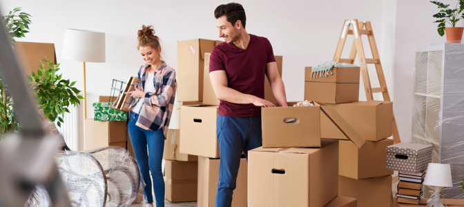 Casal sorridente organizando caixas de papelão em uma sala durante mudança residencial, com ambiente iluminado e móveis embalados
